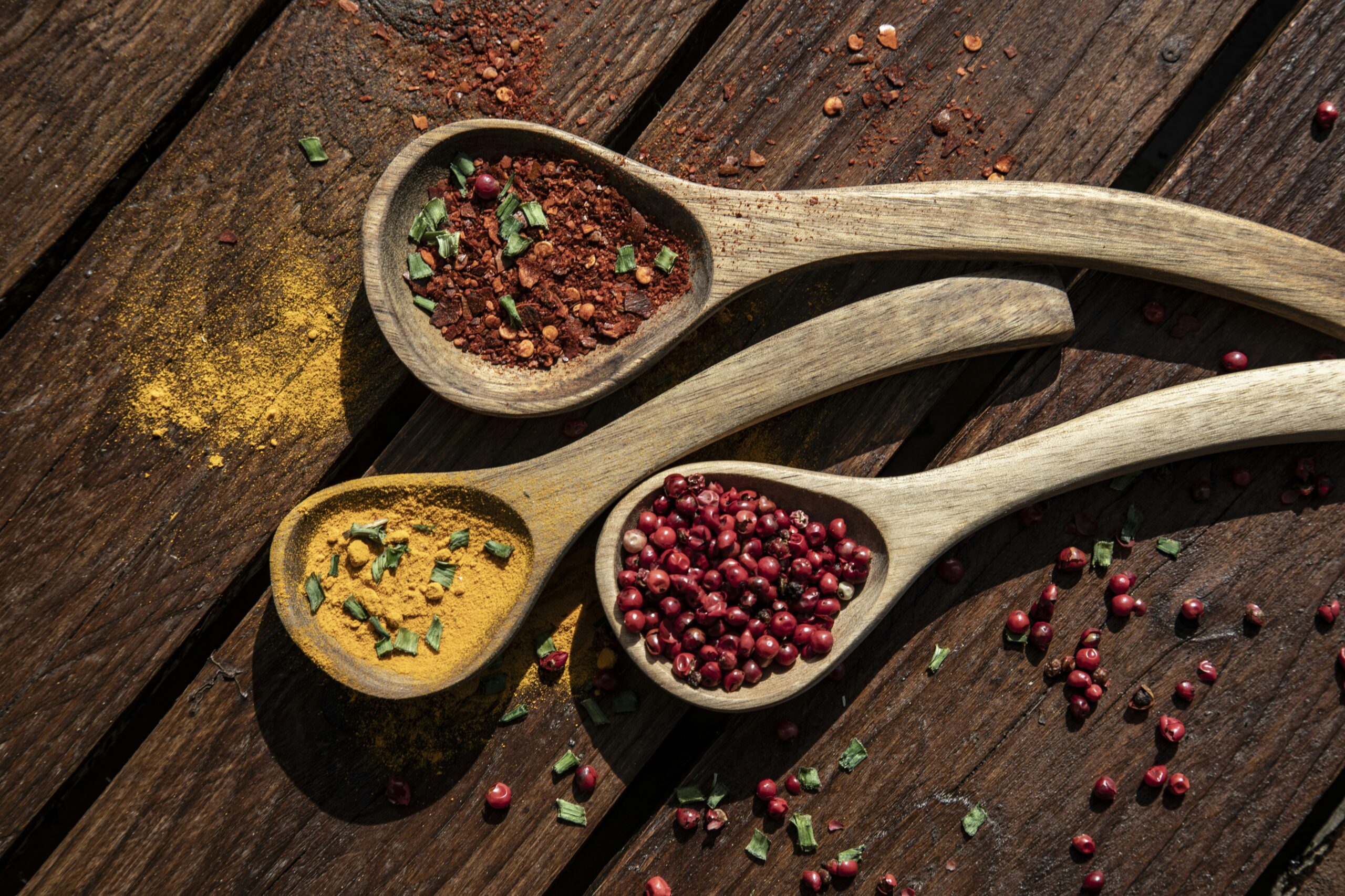 Three wooden spoons containing spices and peppercorns on a wooden surface