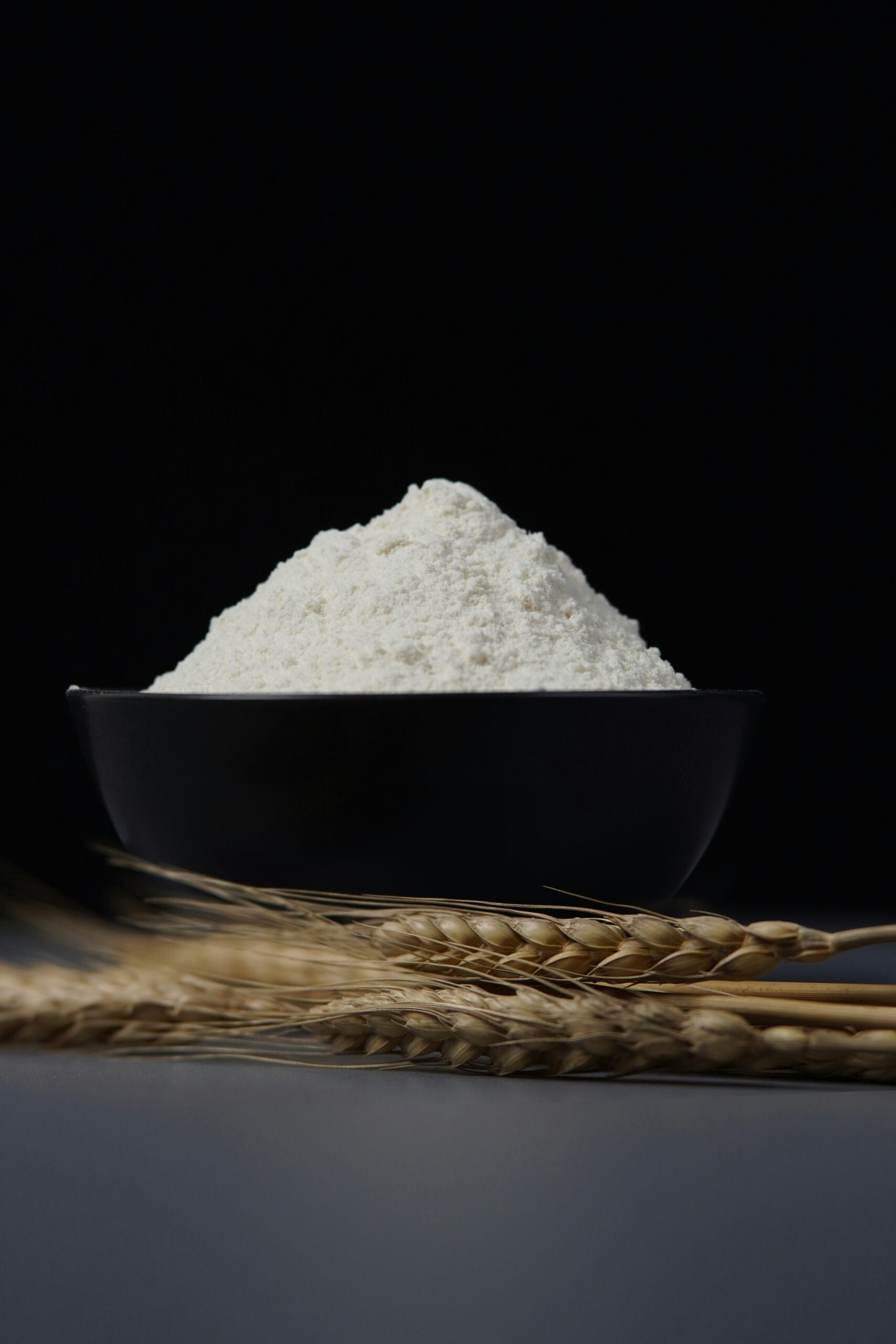 Wheat flour piled in a black bowl with wheat ears beneath